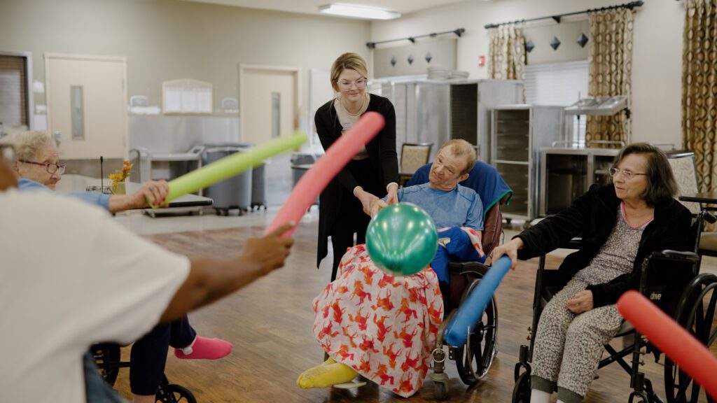 a group of people doing an activity with a pool noodle and balloon at a skilled nursing facility